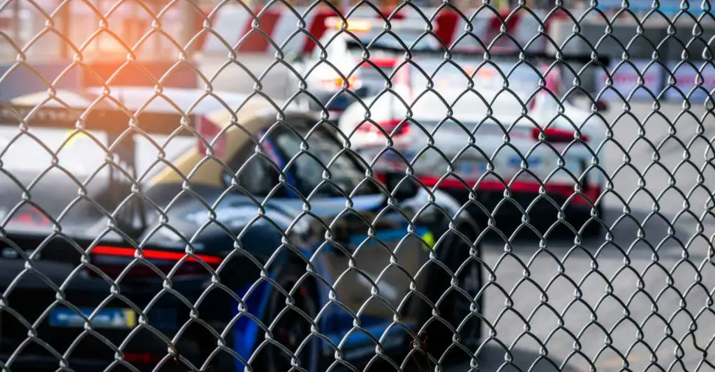 Race cars lined up on track behind chain-link fence, motorsport event.