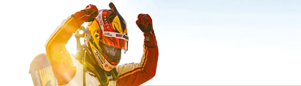 Race car driver celebrating victory with a raised fist, wearing a helmet and racing suit, against a clear sky background.