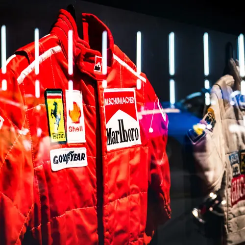 Red racing suit displayed in a museum alongside a white astronaut suit, featuring sponsor logos like Marlboro and Shell, highlighting motorsport and space exploration history.